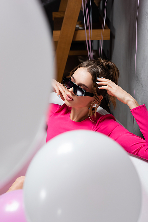 Young woman in sunglasses sitting in bathtub near balloons on blurred foreground