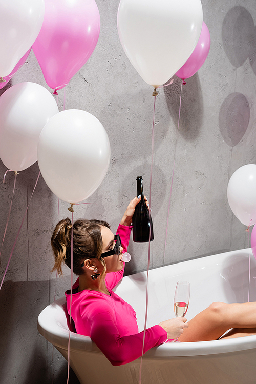 Woman in sunglasses blowing bubblegum and holding champagne in bathtub near balloons