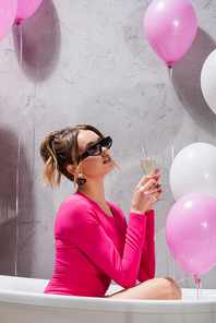 Stylish woman holding glass of champagne in bathtub near balloons