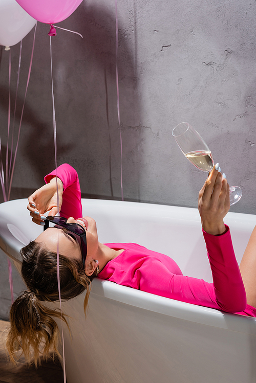 Cheerful woman in sunglasses holding champagne near balloons in bathroom