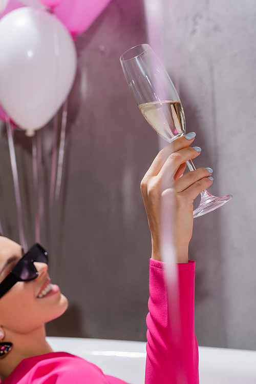 Glass of champagne in hand of smiling woman in sunglasses in bathtub near balloons on blurred background
