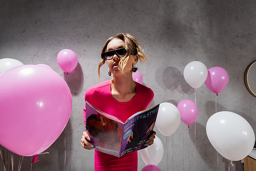 Woman in sunglasses holding fashion magazine near balloons