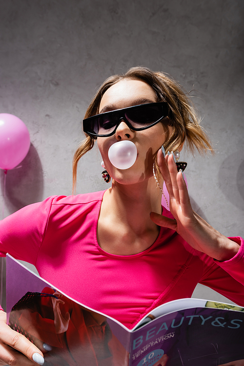 Low angle view of stylish woman with bubblegum holding magazine near blurred balloon