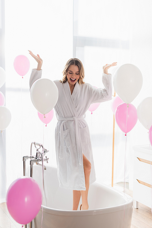 Cheerful woman in bathrobe standing in bathtub near balloons