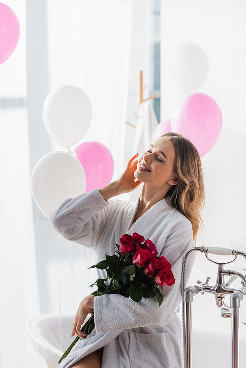 Positive woman in bathrobe holding roses near bathtub and balloons