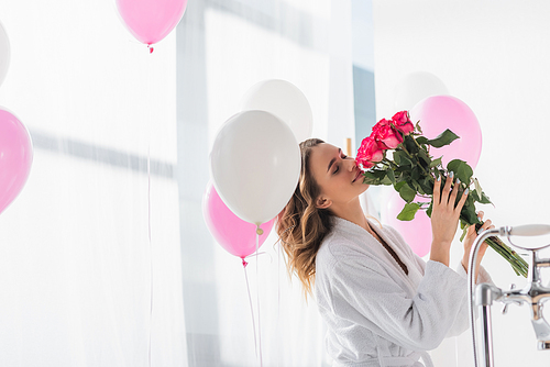 Young woman in bathrobe smelling roses near balloons in bathroom