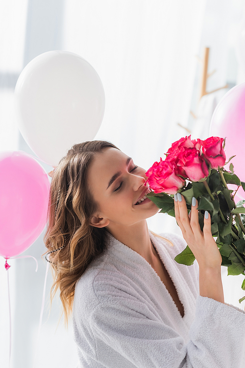 Cheerful woman in bathrobe smelling roses near balloons during birthday celebration