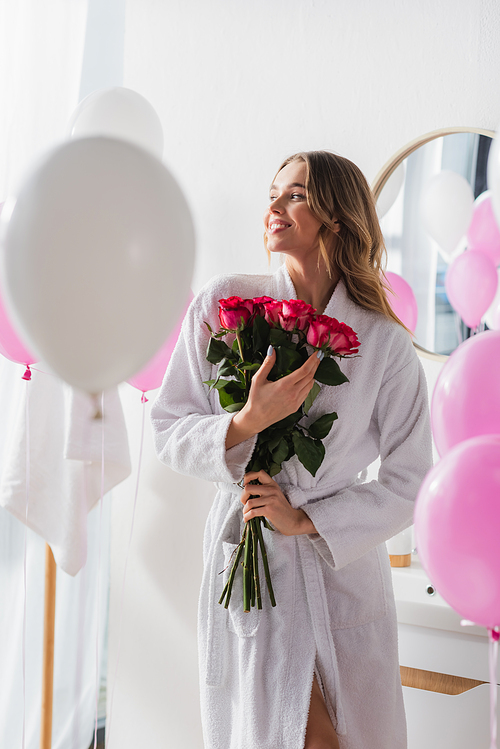 Young woman in bathrobe holding bouquet of roses near balloons in bathroom