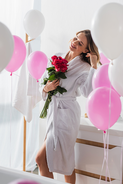 Positive woman in bathrobe holding roses near blurred balloons in bathroom