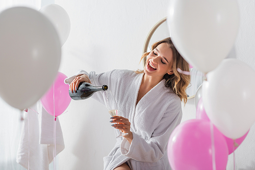 Cheerful woman in bathrobe pouring champagne near balloons in bathroom