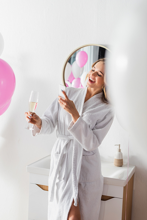 Smiling woman in bathrobe taking photo of champagne near balloons in bathroom