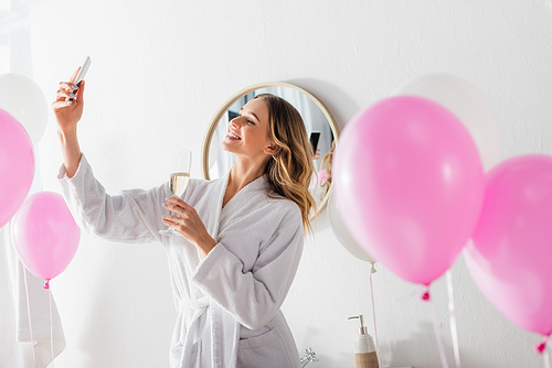 Smiling woman with champagne taking selfie near festive balloons in bathroom