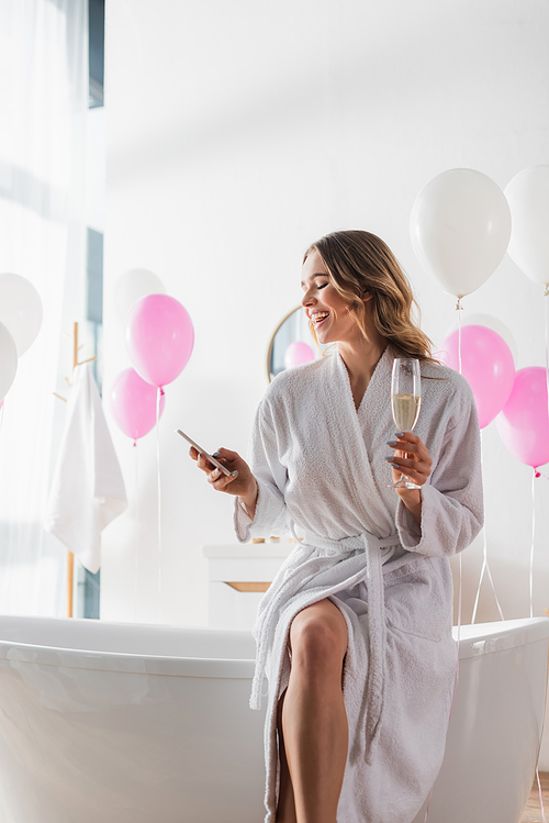 Smiling woman in bathrobe holding champagne and using smartphone near balloons in bathroom