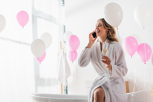 Smiling woman in bathrobe talking on smartphone and holding champagne in bathroom