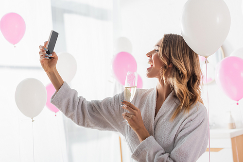 Side view of cheerful woman taking selfie on smartphone and holding champagne near balloons