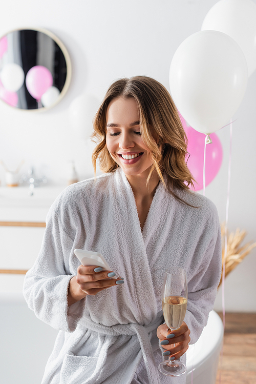 Smiling woman in bathrobe using smartphone and holding champagne in bathroom