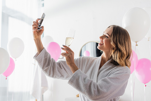 Smiling woman holding champagne and taking selfie with smartphone in bathroom with balloons