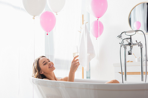 Smiling woman with champagne bathing near balloons in bathroom