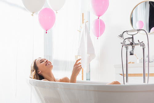 Happy woman holding glass of champagne near balloons in bathroom
