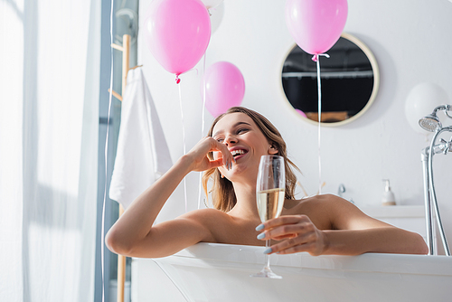 Positive woman with blurred champagne sitting in bathtub near balloons