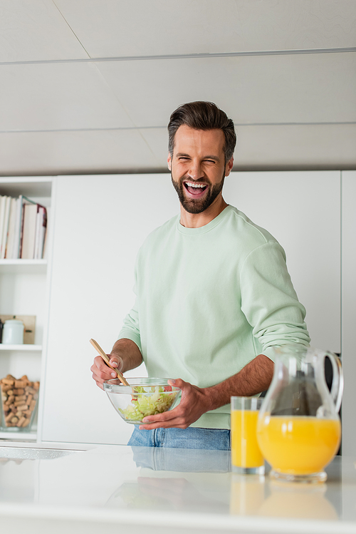 laughing man mixing fresh vegetable salad near orange juice in kitchen