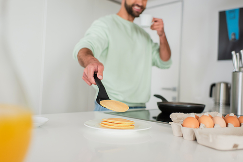 cropped view of smiling blurred man holding coffee cup while cooking pancakes