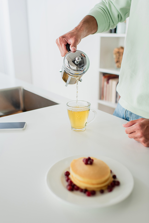 cropped view of man pouring green tea near tasty pancakes on blurred foreground