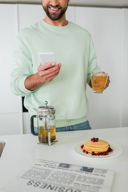 partial view of smiling man holding smartphone and green tea near pancakes in kitchen