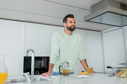 positive man looking away near green tea and delicious pancakes in kitchen