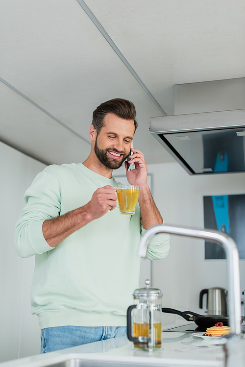 cheerful man with cup of green tea talking on mobile phone in kitchen