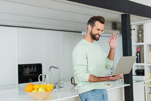 happy man waving hand during video call on laptop in kitchen