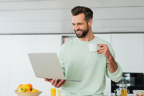 happy man looking at laptop while drinking coffee in kitchen