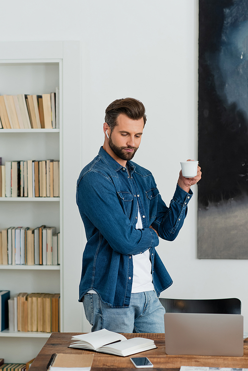 teleworker in denim shirt standing with cup of coffee at workplace