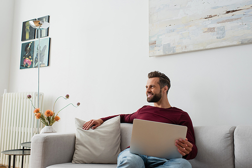 positive man smiling while sitting on sofa at home with laptop