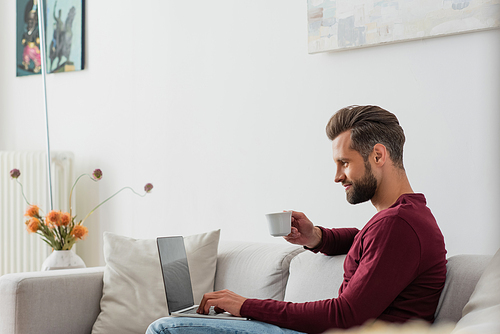 man holding cup of coffee while sitting on sofa with laptop