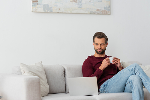 man with cup of coffee sitting on sofa near laptop