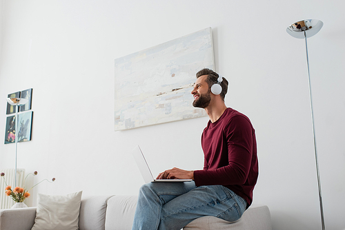 happy man in headphones looking away while sitting on couch with laptop