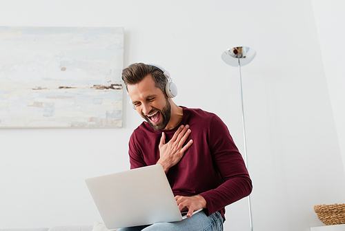 cheerful man in headphones laughing near laptop at home