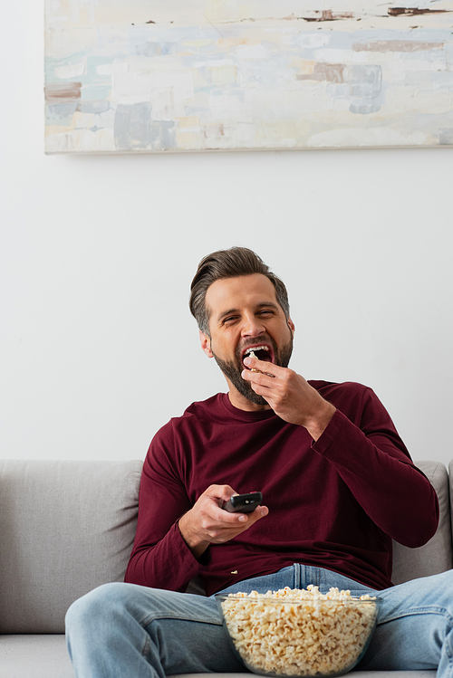 adult man holding remote controller and eating popcorn on couch