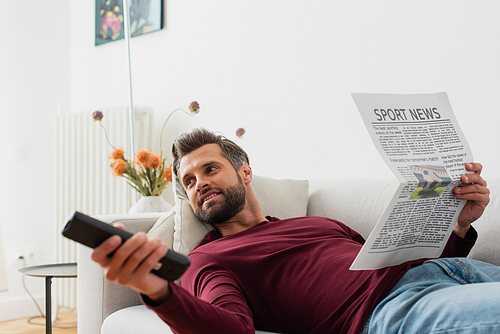 joyful man clicking tv channels while lying on couch with newspaper
