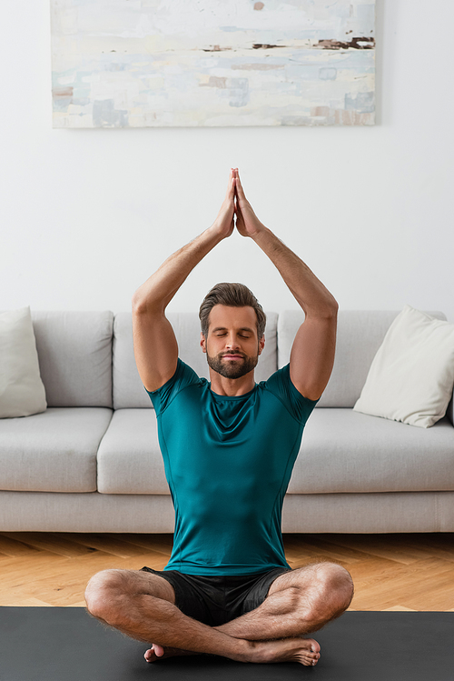 man with praying hands and closed eyes meditating in lotus pose at home