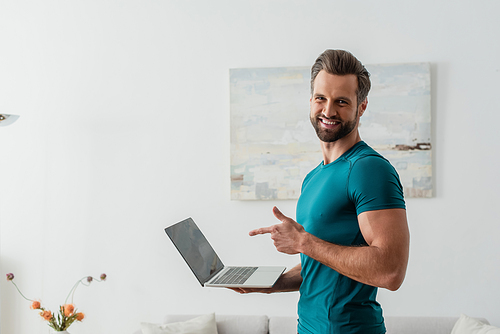 happy man pointing with finger at laptop while smiling at camera