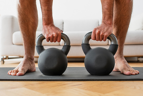 partial view of barefoot man lifting kettlebells on fitness mat