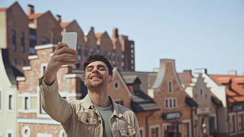 cheerful man taking selfie with blurred buildings on background