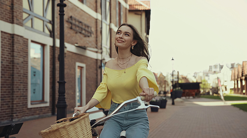 dreamy young woman in yellow blouse smiling while riding on bicycle outside