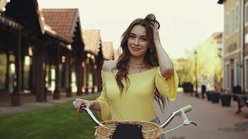 happy young woman in yellow blouse adjusting hair while riding on bicycle outside