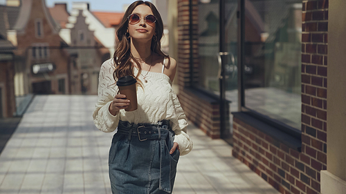 stylish young woman in sunglasses holding coffee to go and walking outside