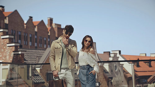 trendy couple in sunglasses posing with blurred buildings on background