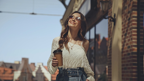 smiling young woman in sunglasses holding coffee to go outside