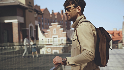 side view of stylish man in sunglasses with backpack standing outside
